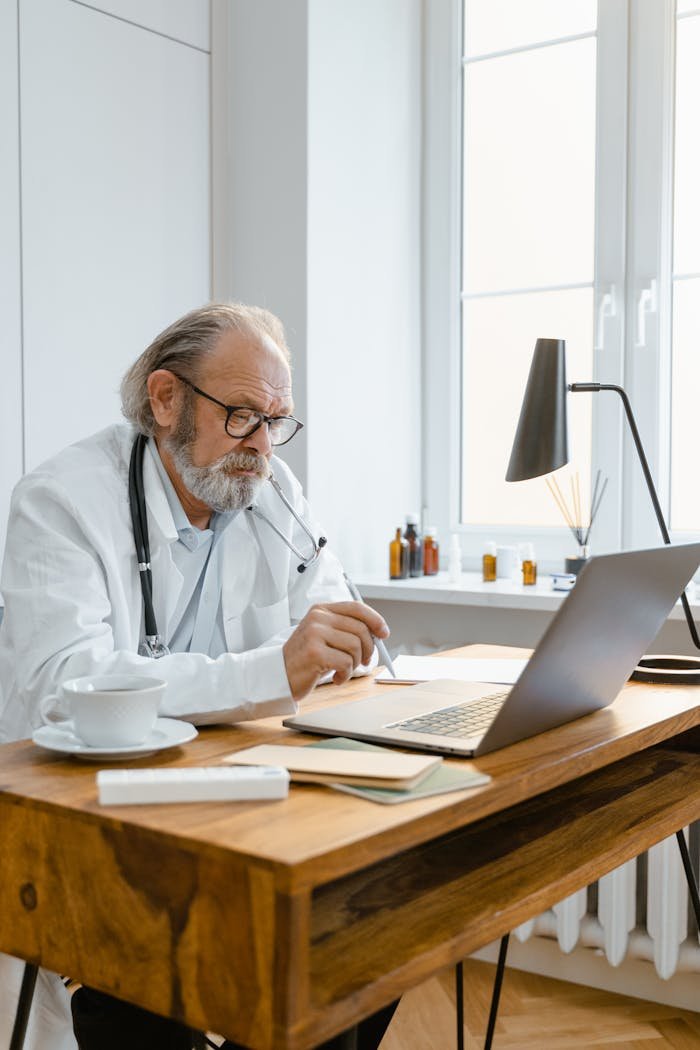 Elderly doctor in office using laptop for telemedicine consultation, showcasing modern healthcare tech.